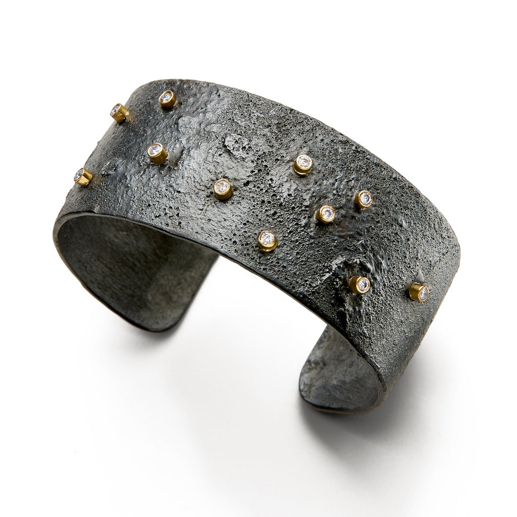 The Floating in the Dark cuff bracelet in blackened sterling silver, adorned with small diamonds set in 18k yellow gold bezels, makes a bold statement while being suitable for everyday wear. The blackened sterling silver provides a striking contrast to the sparkle of the diamonds and the warmth of the yellow gold, creating a piece that is both eye-catching and versatile.  • Blackened sterling silver, 8k yellow gold bezels, diamonds ct: 0.33  • Height: 1"